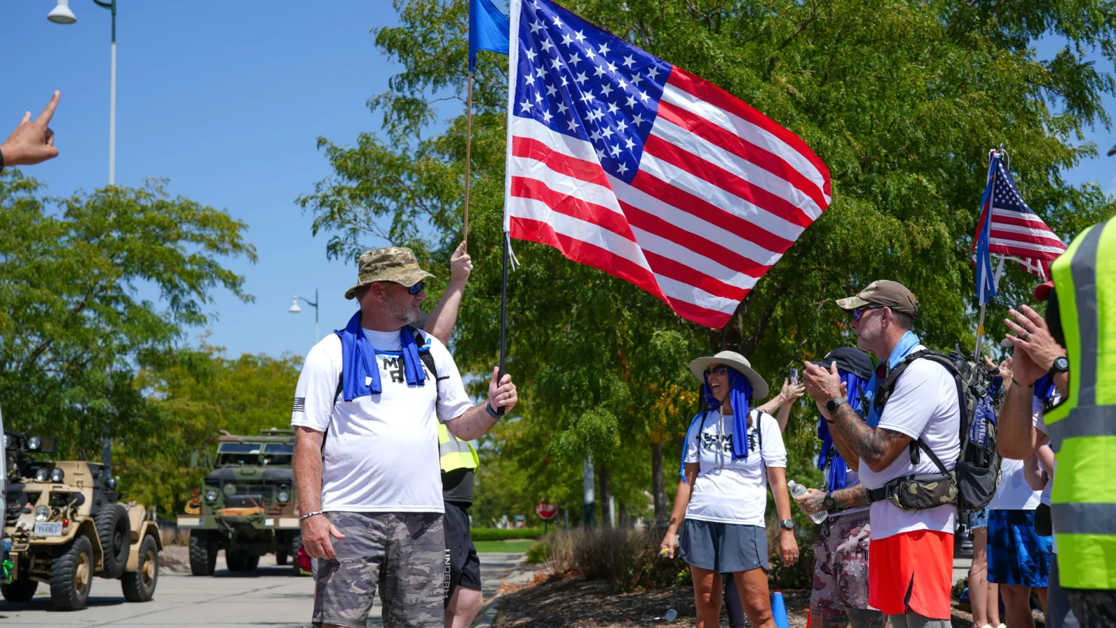 A 50 Mile March participant carrying the American flag alongside fellow marchers and a military vehicle.